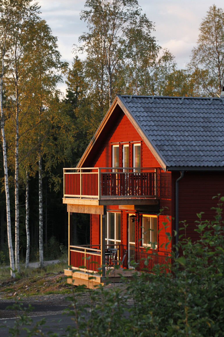 Röd stuga omgiven av skog i kvällsljus.