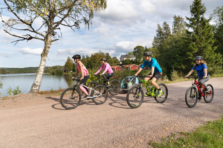 Familj cyklar på grusväg vid sjö.