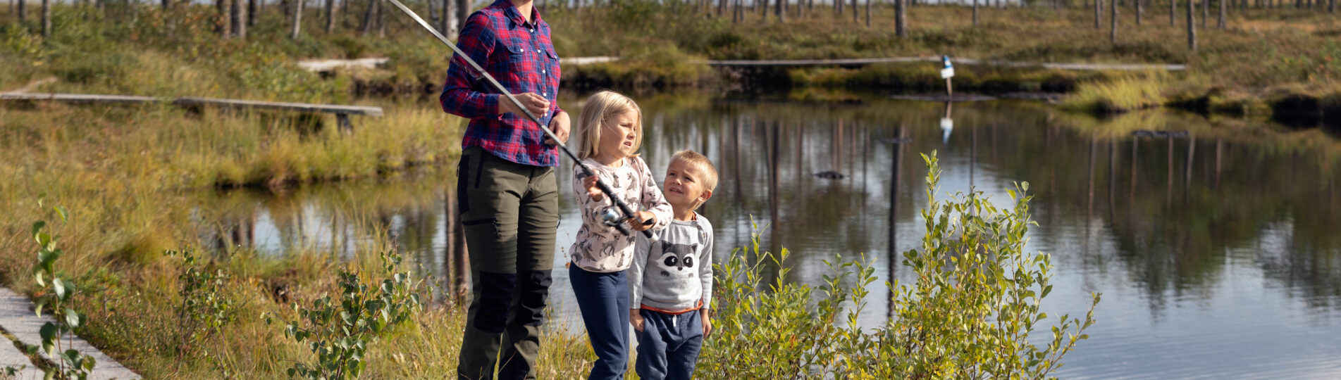 Mamma fiskar med två barn vid tjärn.