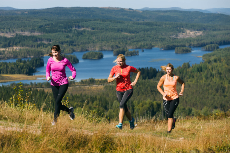 Tre kvinnor springer terränglöpning uppför berget med utsikt över Säfssjön och skog.