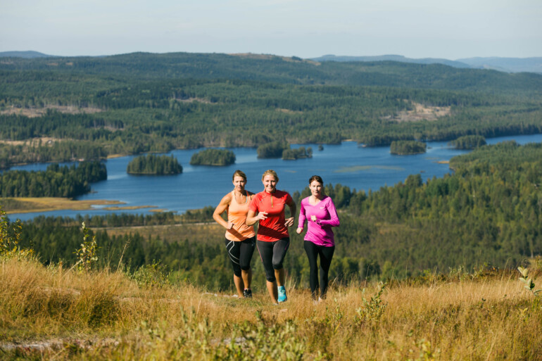 Tre kvinnor springer uppför ett berg med utsikt över skog och sjö.
