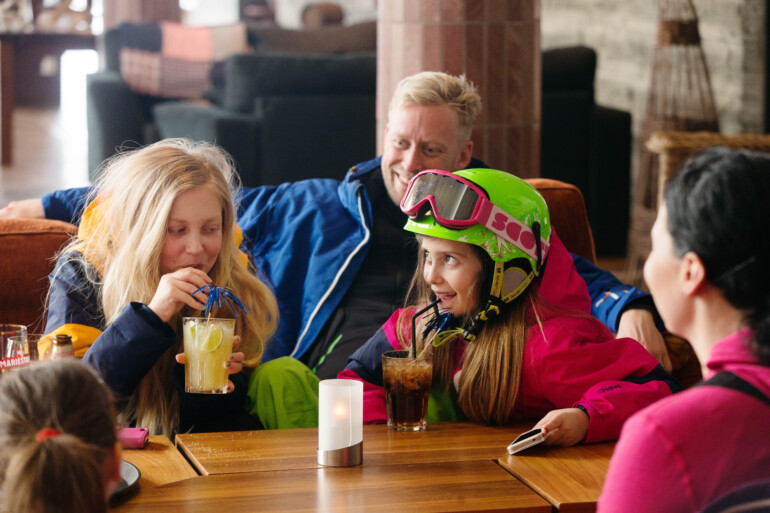 Familj i skidkläder sitter i soffa och dricker läsk.