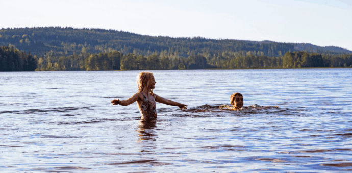 2 barn som badar i sjön, skog och blå himmel i bakgrunden.