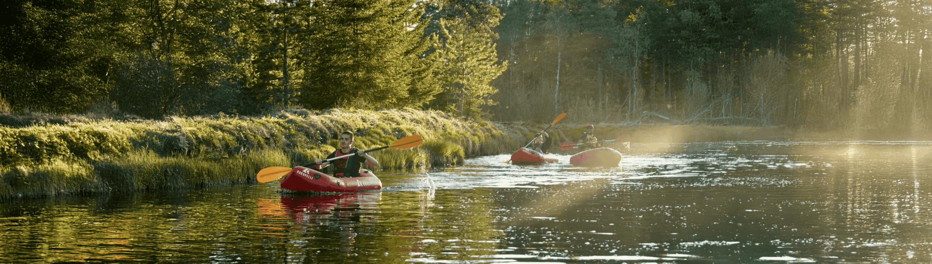 tre personer paddlar i varsin uppblåsbar packraft i solnedgången. Stilla vatten och grön skog i bakgrunden