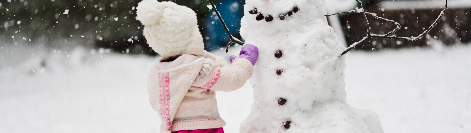 A toddler girl in a pink snowsuit with wooly hat toque builds a large snowman with carrot nose and smiling face in backyard with snowflakes snow falling in winter at Christmas